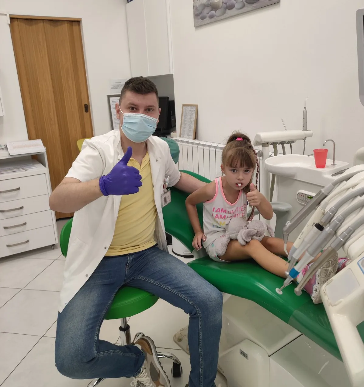 Djevojčica sa svojom plišanom igračkom u stolici, uz dr. Asifa — oboje pokazuju palac gore